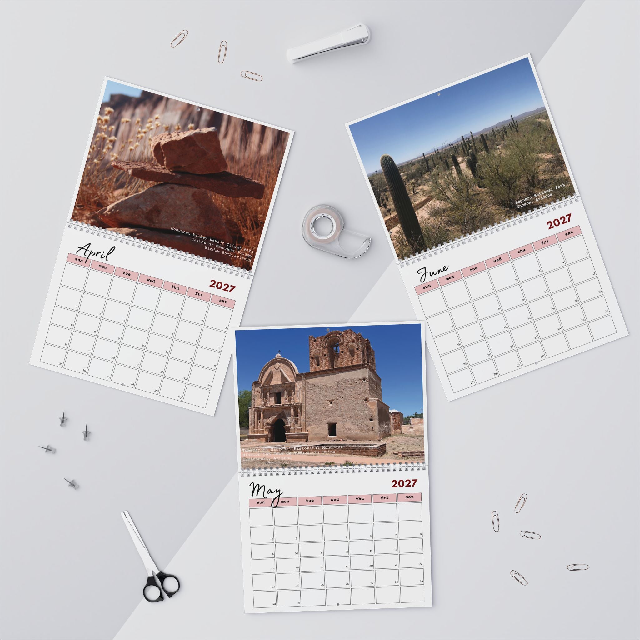 Three spiral-bound monthly calendar pages with desert landscape photos are arranged on a light gray surface with small office props like paper clips, tape, scissors, and a pen.