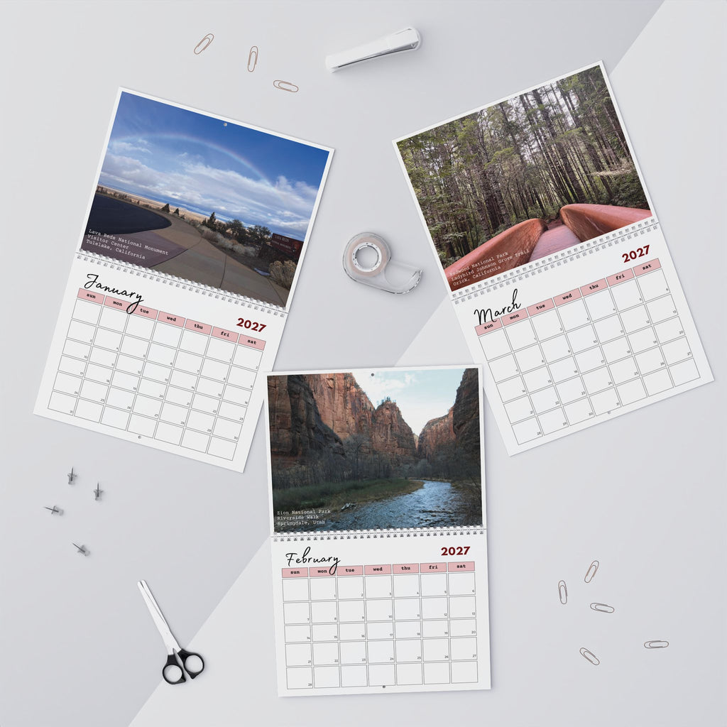 Three spiral-bound monthly calendar pages showing January, March, and February 2027 with landscape photographs, arranged on a light gray background with office props like paper clips, tape, scissors, and a pen.