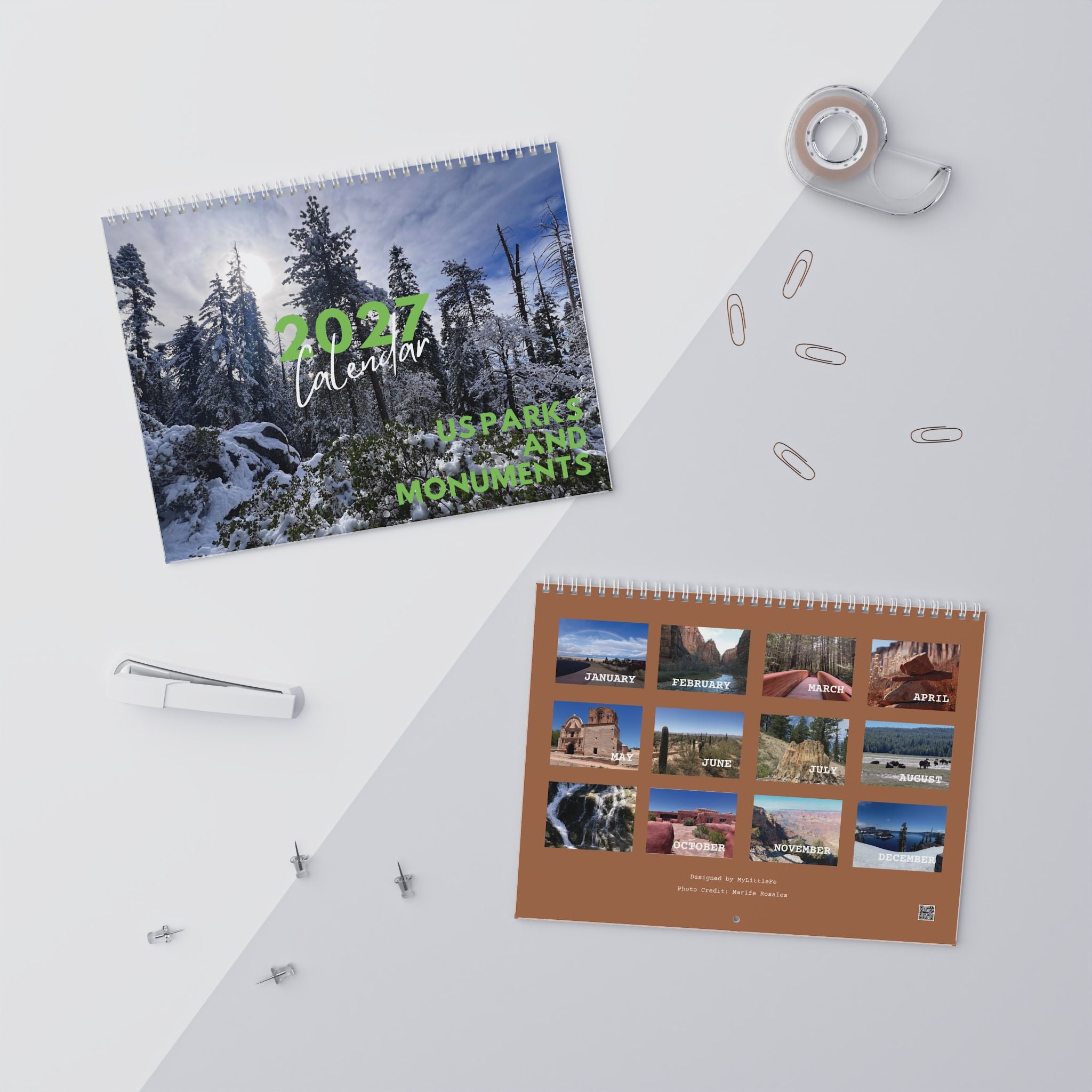 Two spiral-bound wall calendars displayed on a light gray surface with desk props. The larger calendar on the left shows a snowy forest scene with green text reading '2027 Calendar' and 'APPAREL AND MONUMENTS.