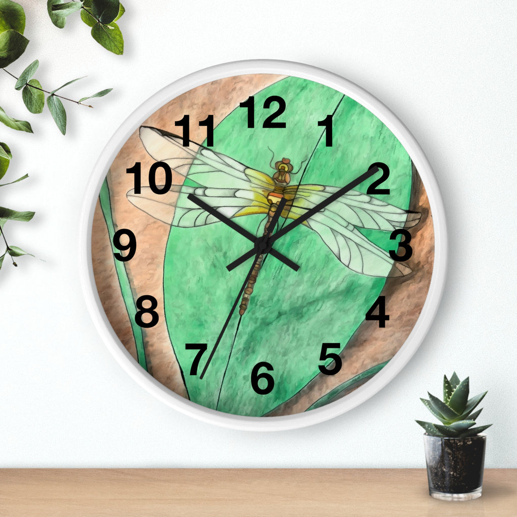 Wall clock with dragonfly design on a green background, placed on a wooden surface with plants.