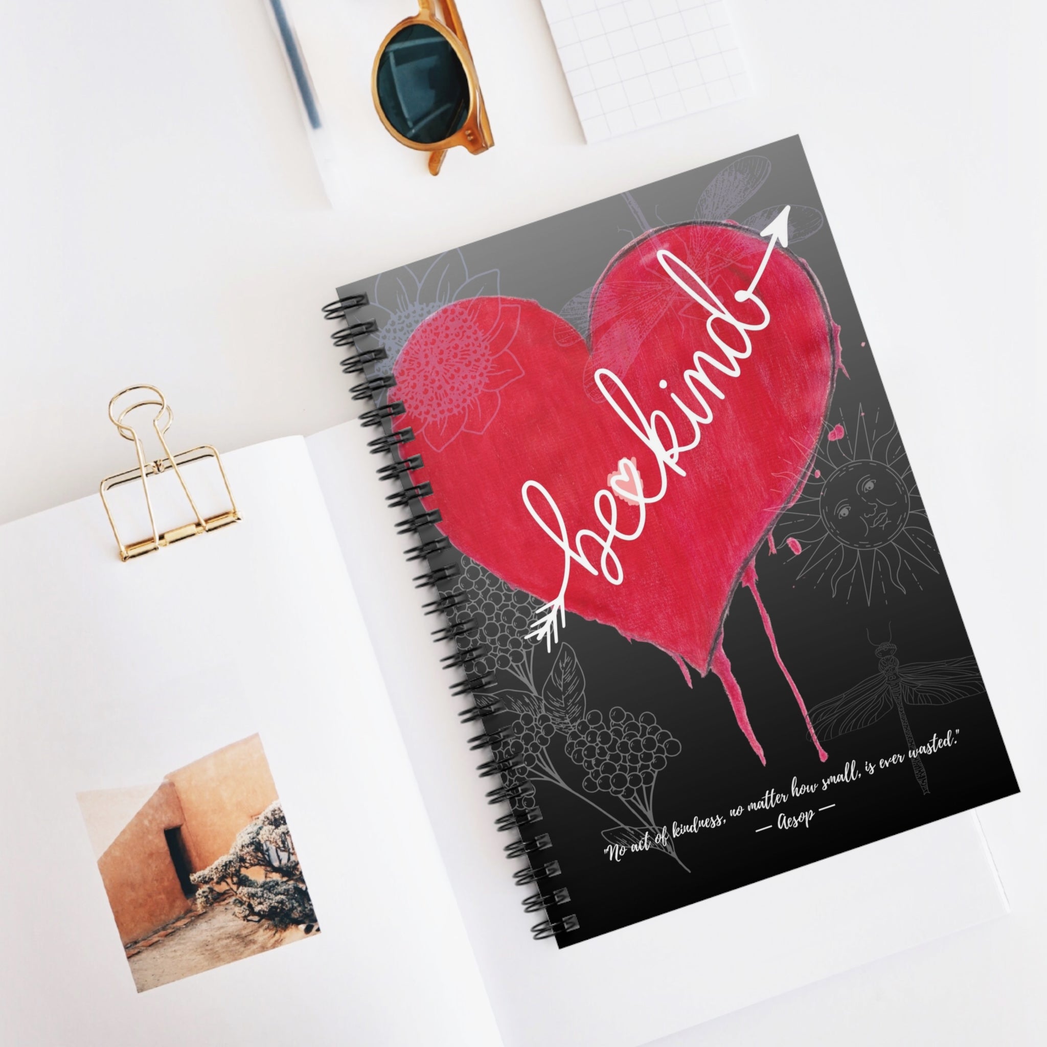Notebook with a red heart and 'be kind' text on a white surface with a photo and clip.