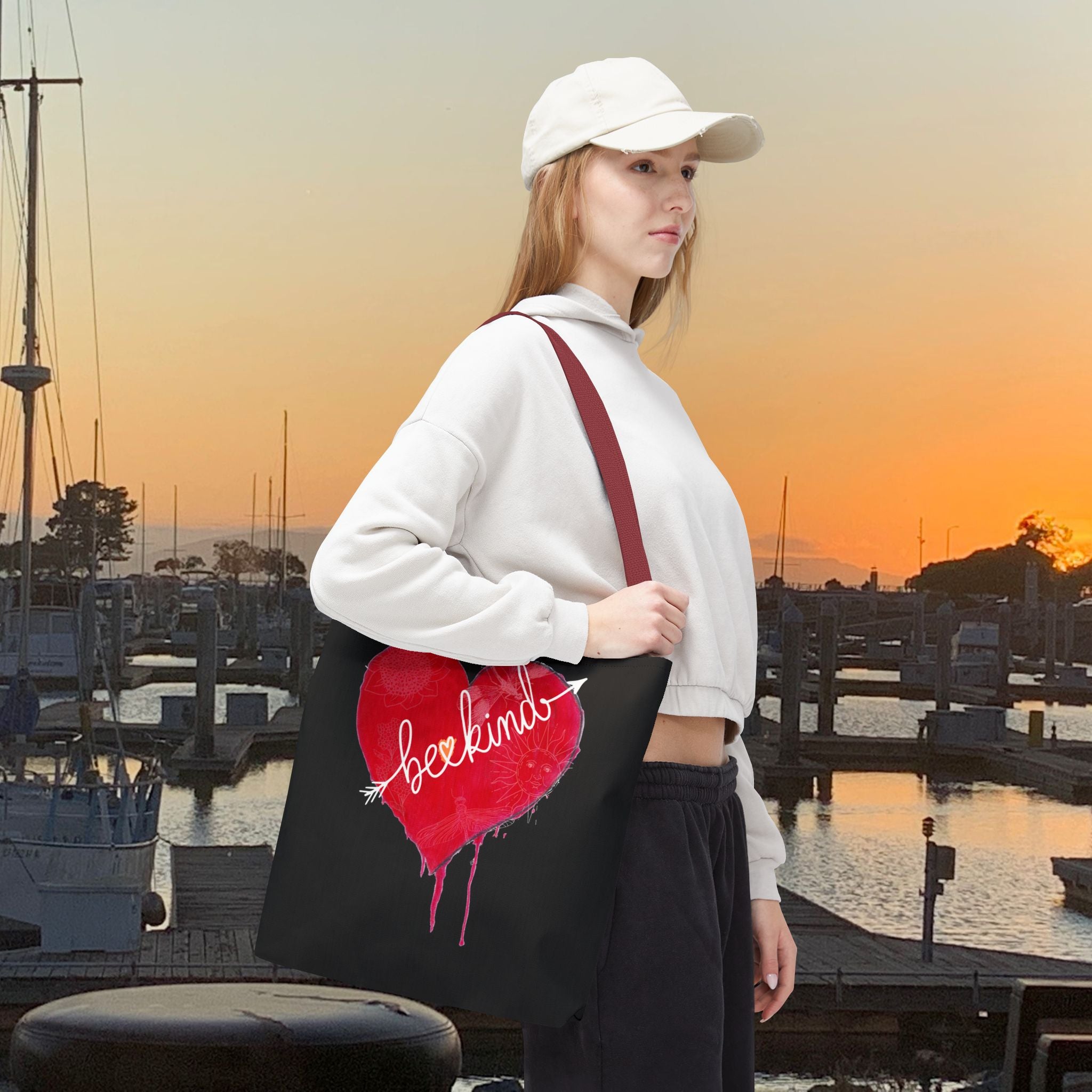 Woman holding a tote bag with a red heart design at a marina during sunset.
