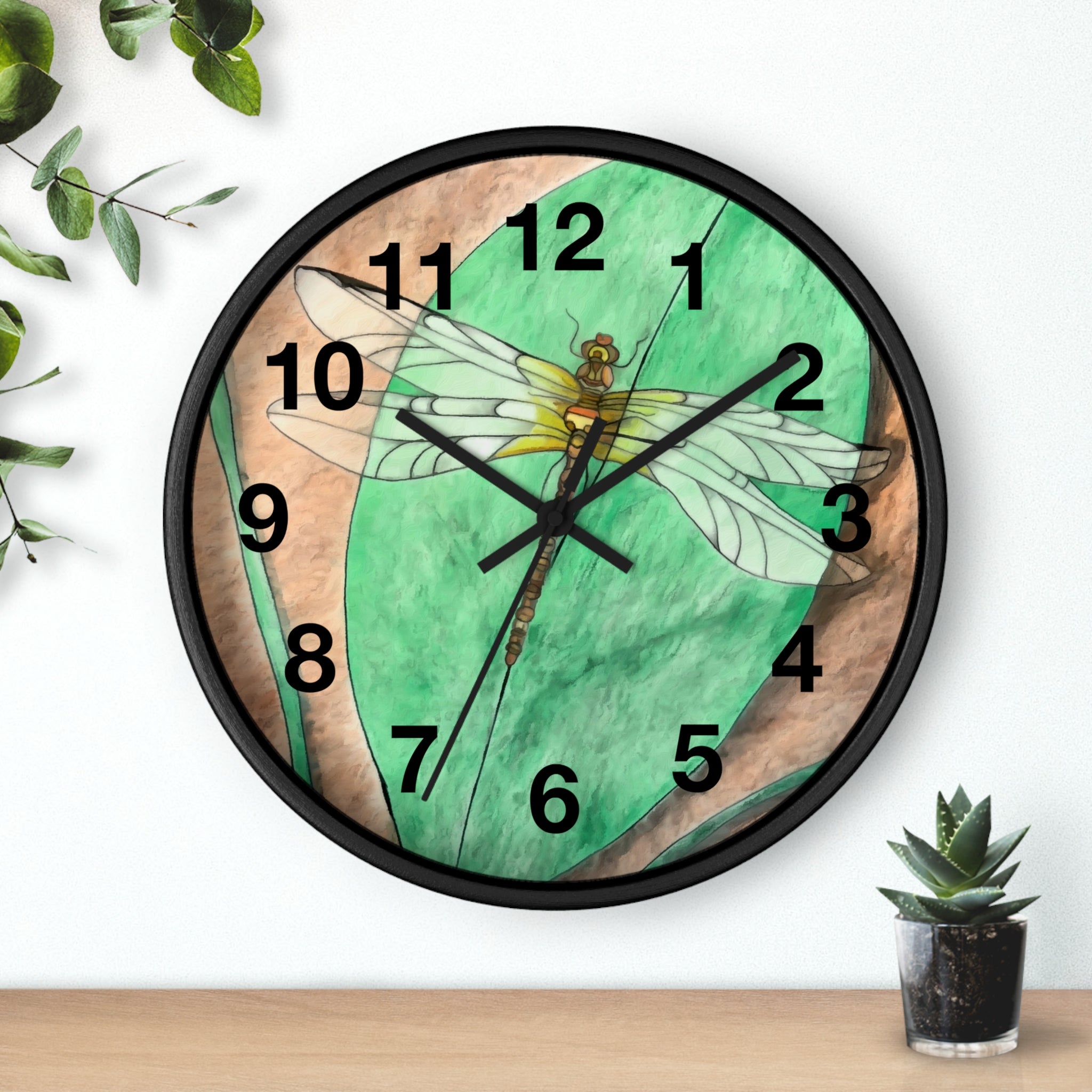 Wall clock with dragonfly design on a green and brown background, placed on a wooden surface with a plant.