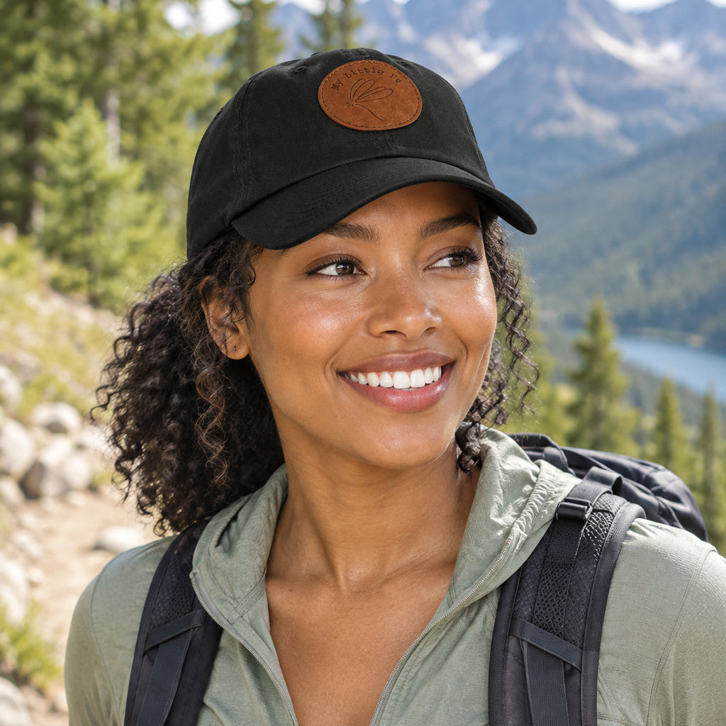 Person hiking wearing a black cap with My Little Fe and Dragonfly stick drawing logo on a brown patch 