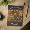Notebook with spider illustration and 'Web Developer at Work' text on a wooden surface with stationery items.
