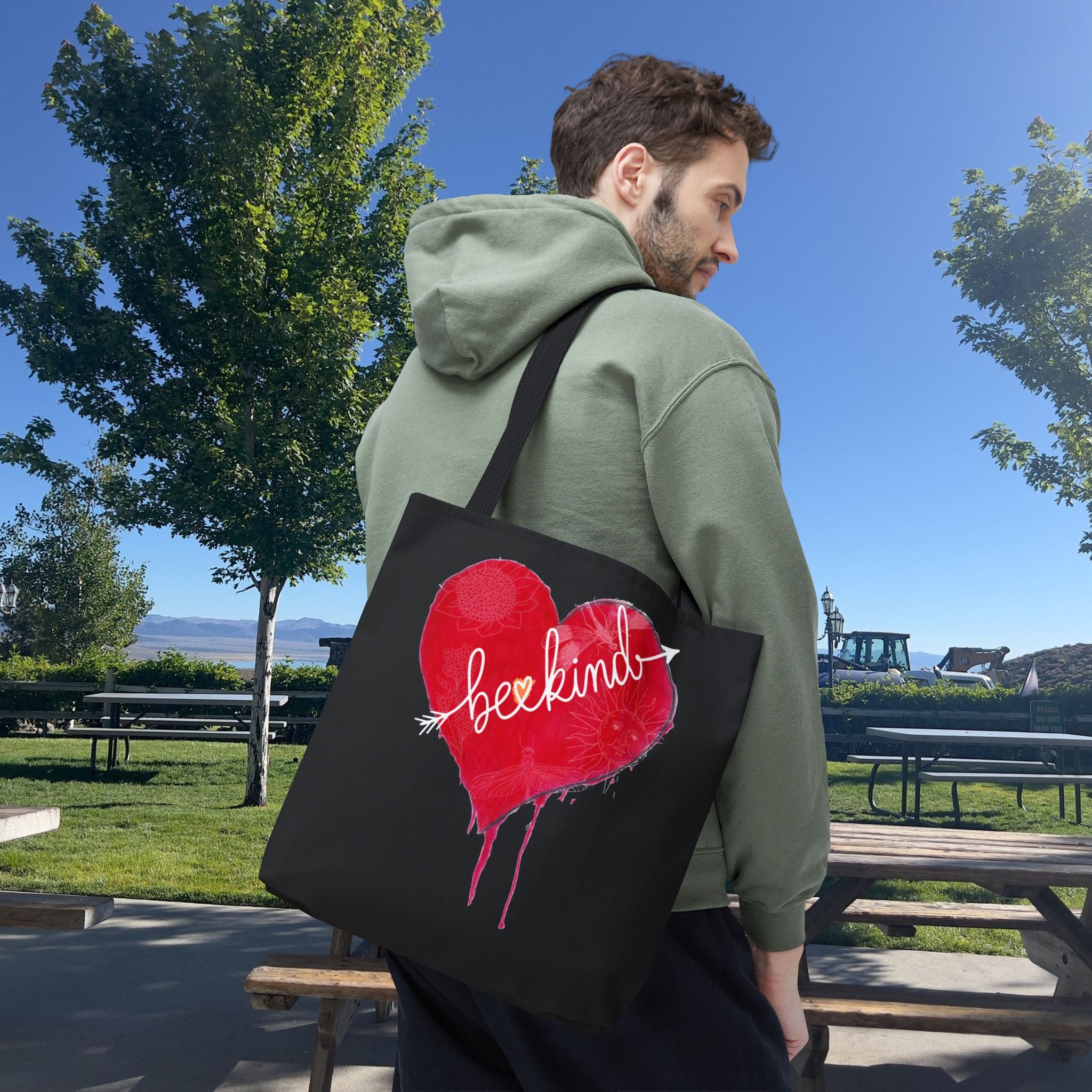 Person wearing a green hoodie with a black tote bag featuring a red heart and 'be kind' text in a park.