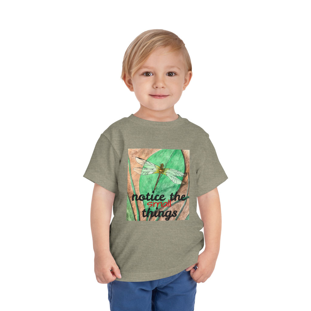 A young child wearing a heather olive green t-shirt with a square graphic print of a dragonfly on a green leaf and the text 'notice the things'. The child is also wearing blue jeans, photographed against a solid white background.