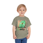 A young child wearing a heather olive green t-shirt with a square graphic print of a dragonfly on a green leaf and the text 'notice the things'. The child is also wearing blue jeans, photographed against a solid white background.