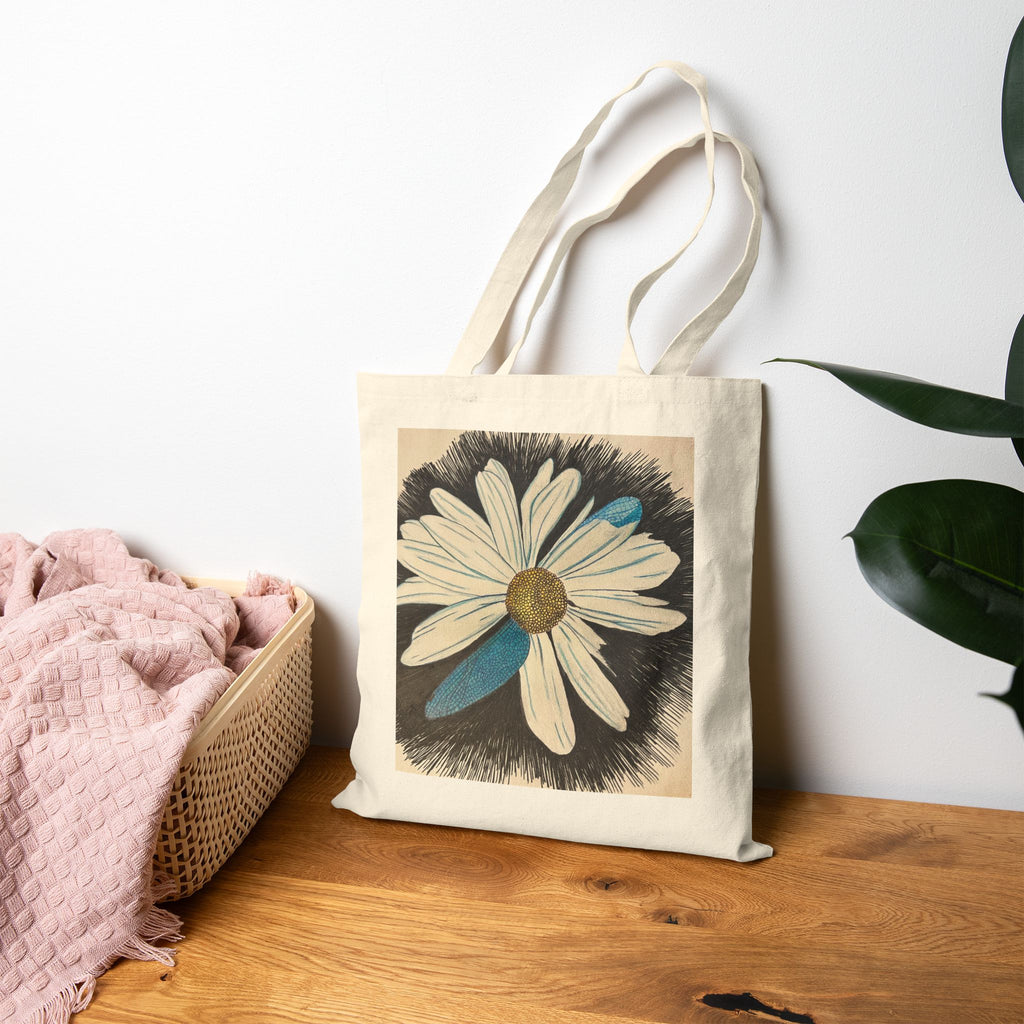 Tote bag with a floral design on a wooden surface next to a pink blanket and plant leaves.