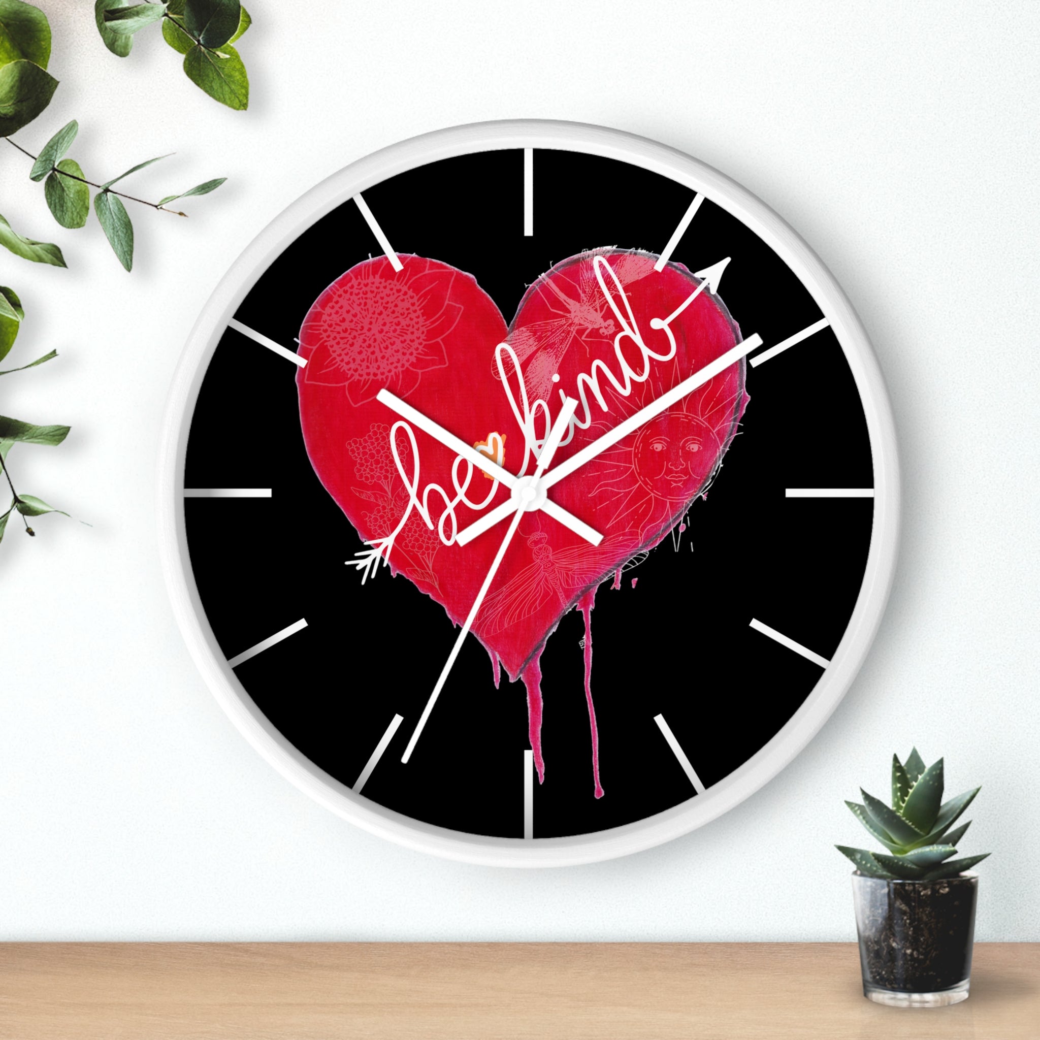 Wall clock with a red heart and 'be kind' text on a black background, placed on a wooden surface with plants.