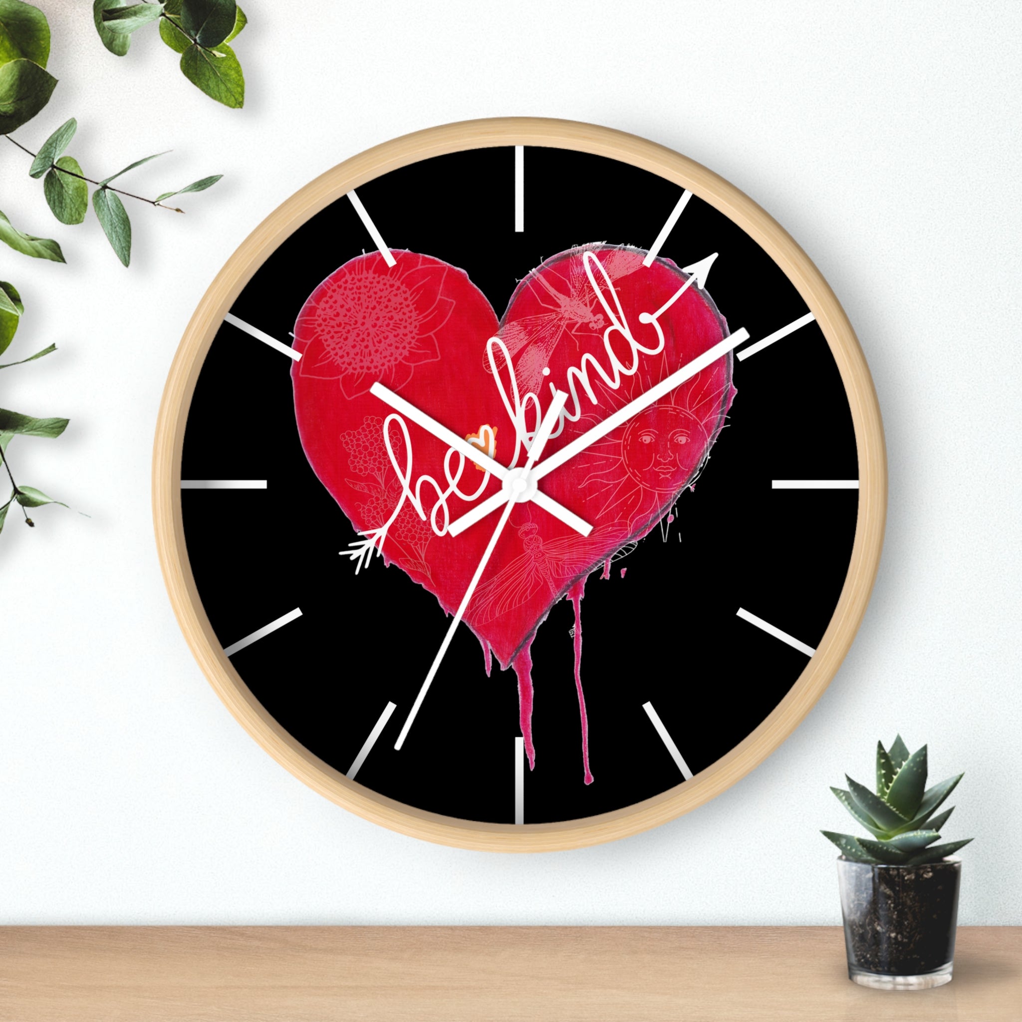 Wall clock with a red heart and 'be kind' text on a black background, placed on a wooden surface with a plant.