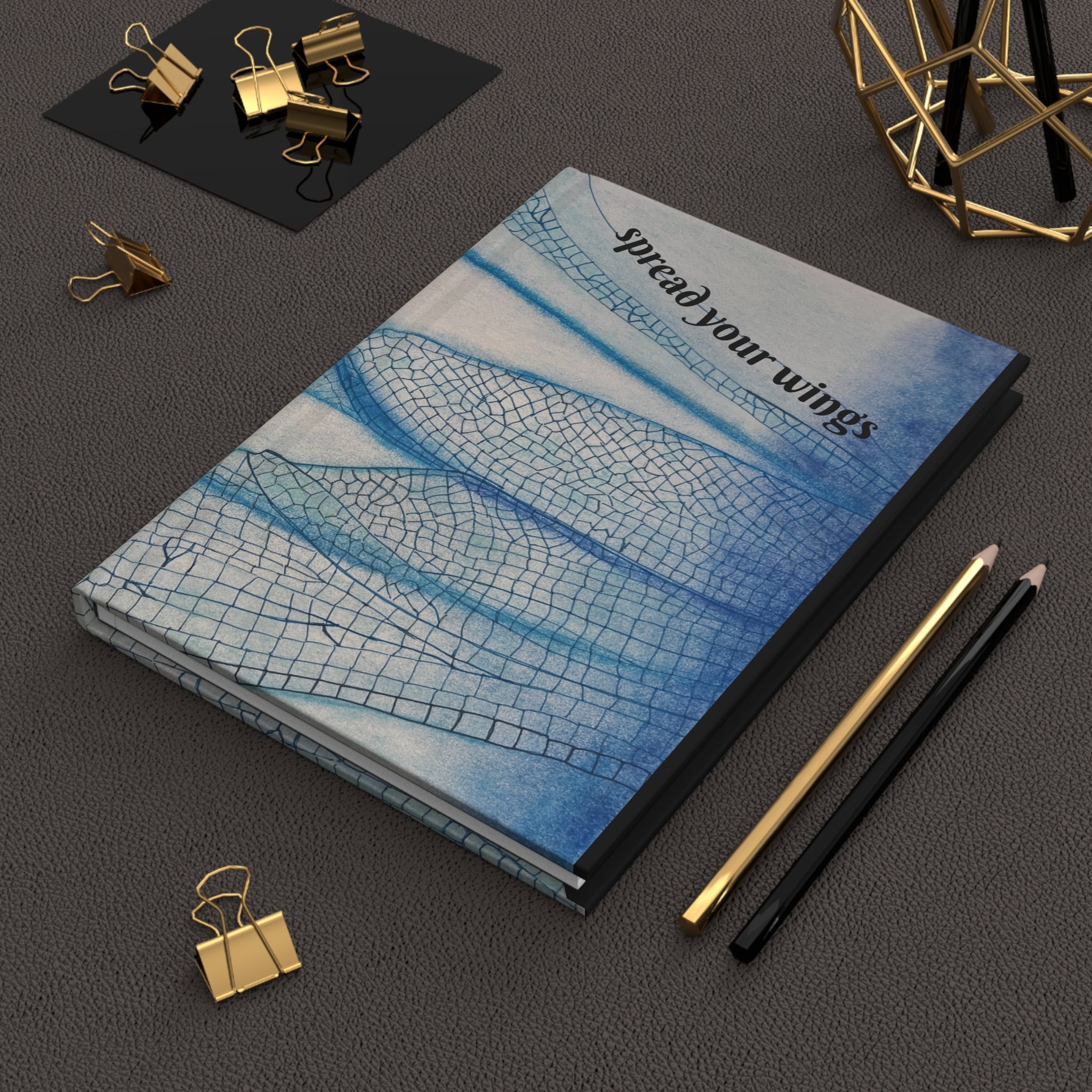 Notebook with 'spread your wings' text on a dark surface with office supplies.