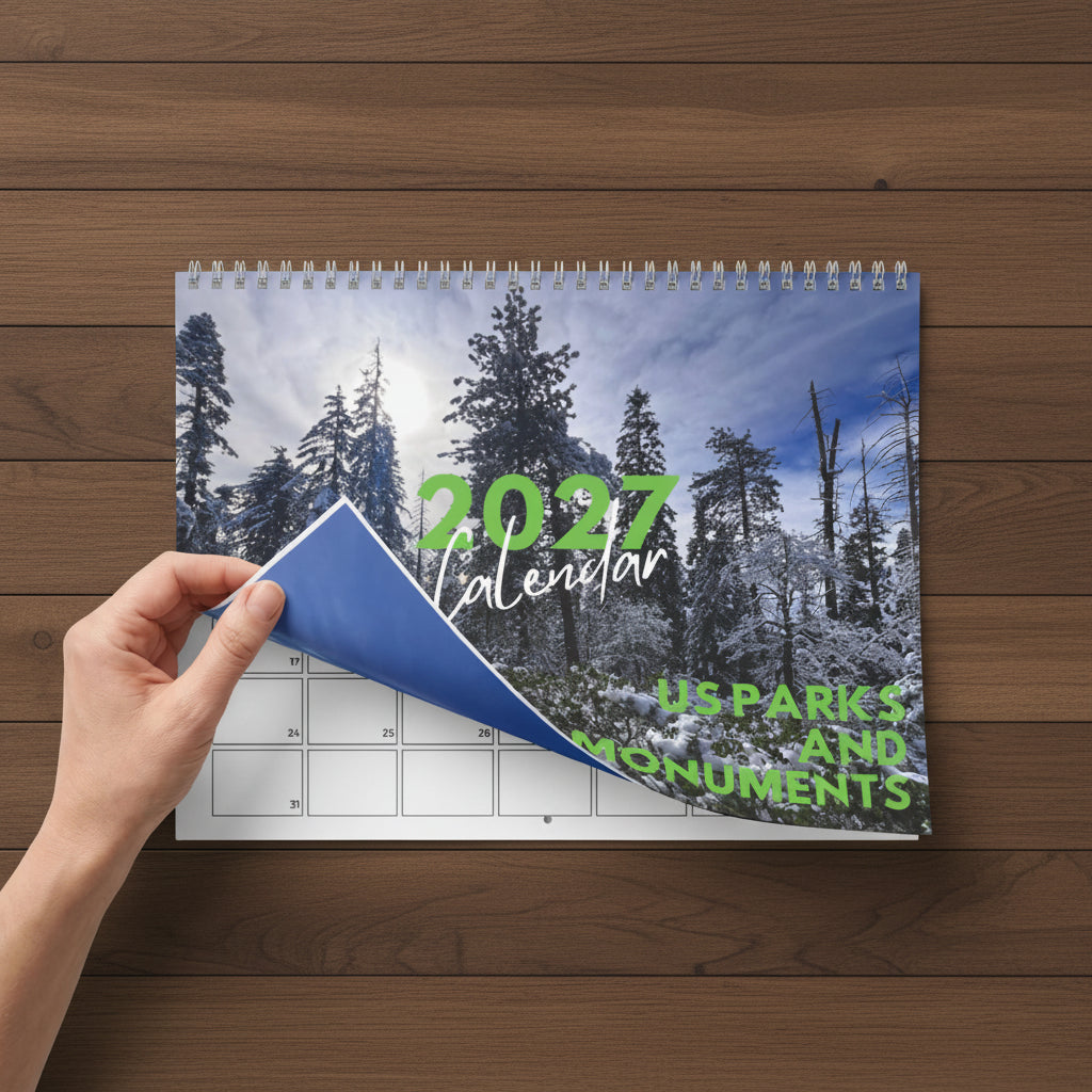 A spiral-bound 2027 wall calendar being held down by a hand, showing a snowy pine forest cover with green text reading '2027 Calendar' and 'US Parks and Monuments'.