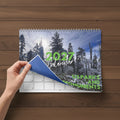 A spiral-bound 2027 wall calendar being held down by a hand, showing a snowy pine forest cover with green text reading '2027 Calendar' and 'US Parks and Monuments'.