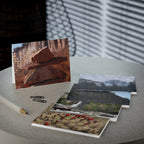 A set of landscape photography postcards arranged on a light gray tabletop, with one card standing upright showing stacked rocks in warm brown and orange tones. Other cards fanned out below show snowy mountains, a waterfall, and a field of red flowers.
