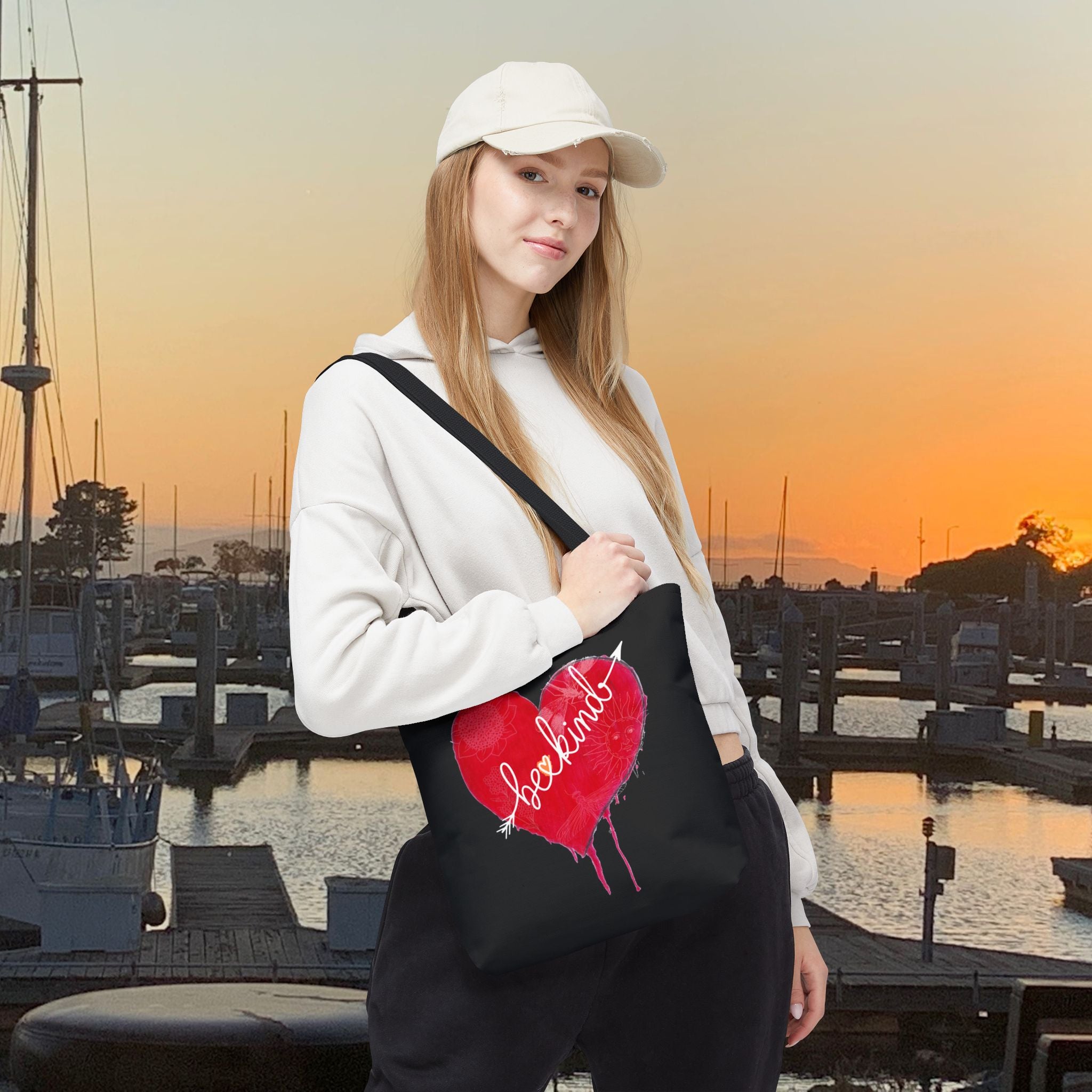 Woman holding a black tote bag with a red heart design at sunset by a marina.