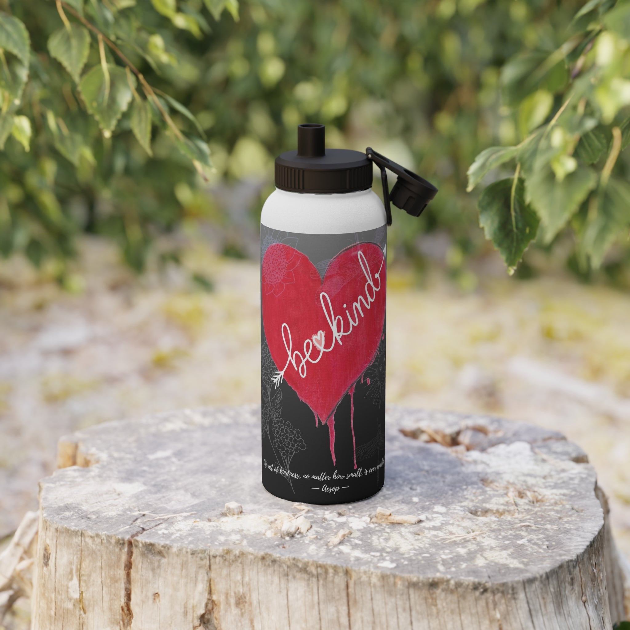 Water bottle with a red heart and 'be kind' text on a wooden stump with greenery in the background