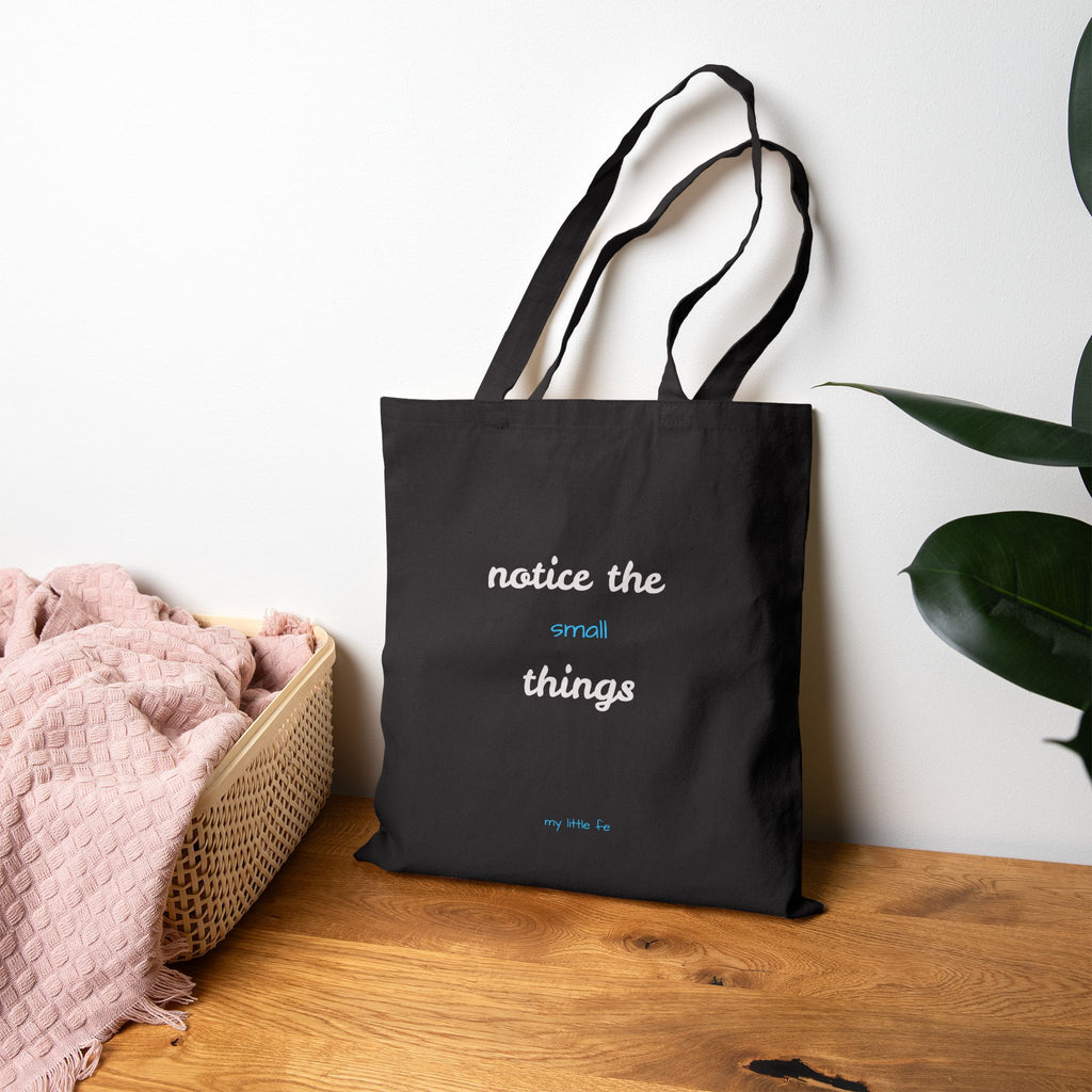 Black tote bag with 'notice the small things' text on a wooden surface with a plant in the background