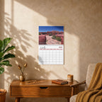A wall-mounted 2023 desk calendar showing a desert landscape photograph, styled in a warm, sunlit room on a wooden console table next to a chair with a brown throw blanket.