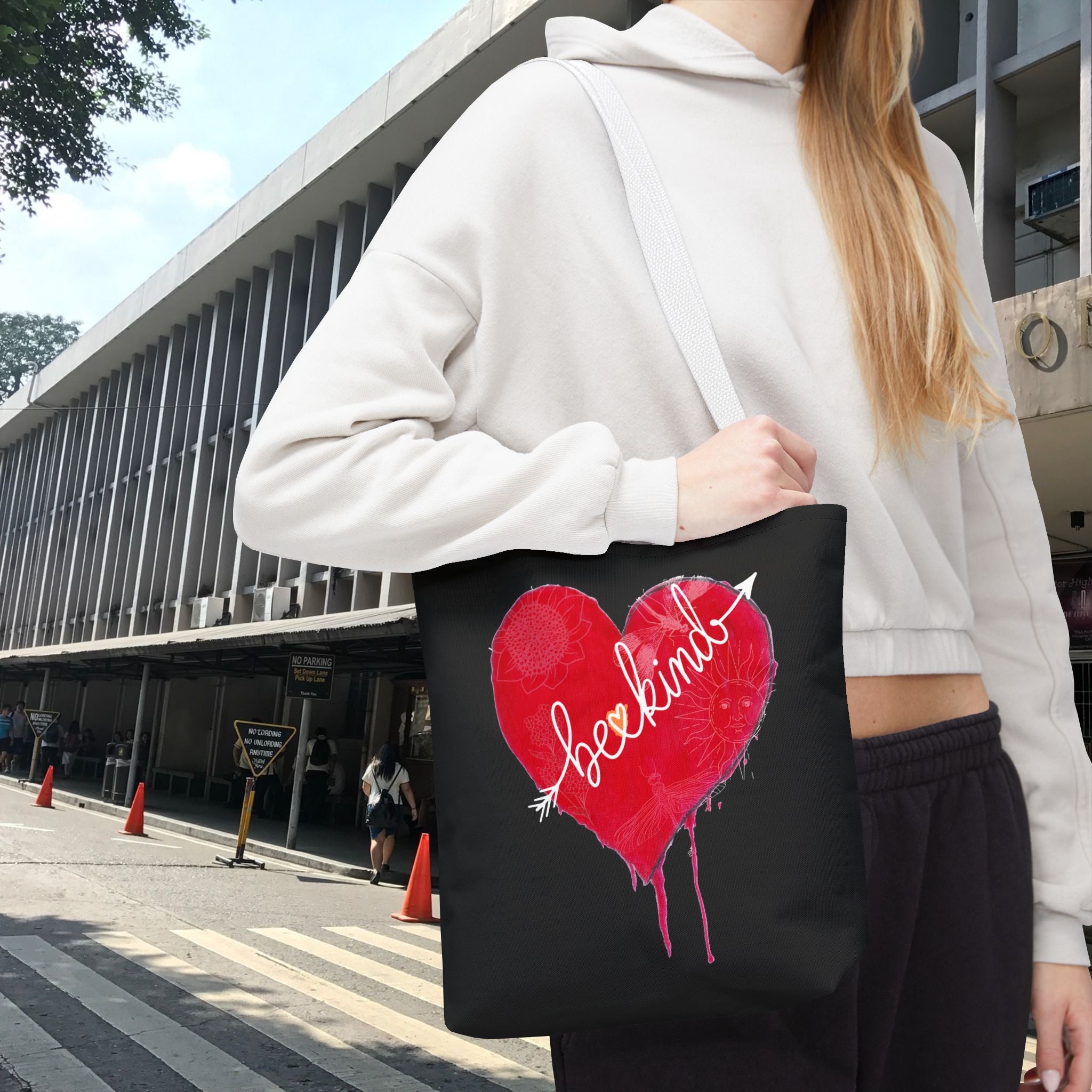 Person holding a black tote bag with a red heart design outdoors.