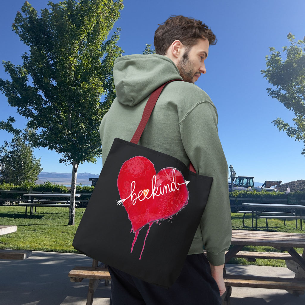Person holding a black tote bag with a red heart and 'be kind' text in a park setting.