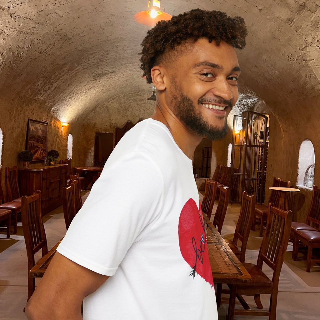 Man in a white t-shirt with a red heart design standing in a rustic room with wooden tables and chairs.