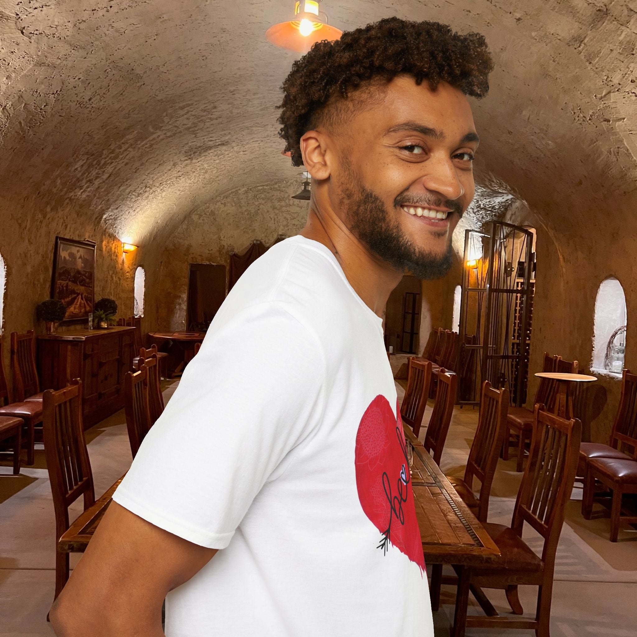 Man in a white t-shirt with a red heart design standing in a rustic room with wooden tables and chairs.