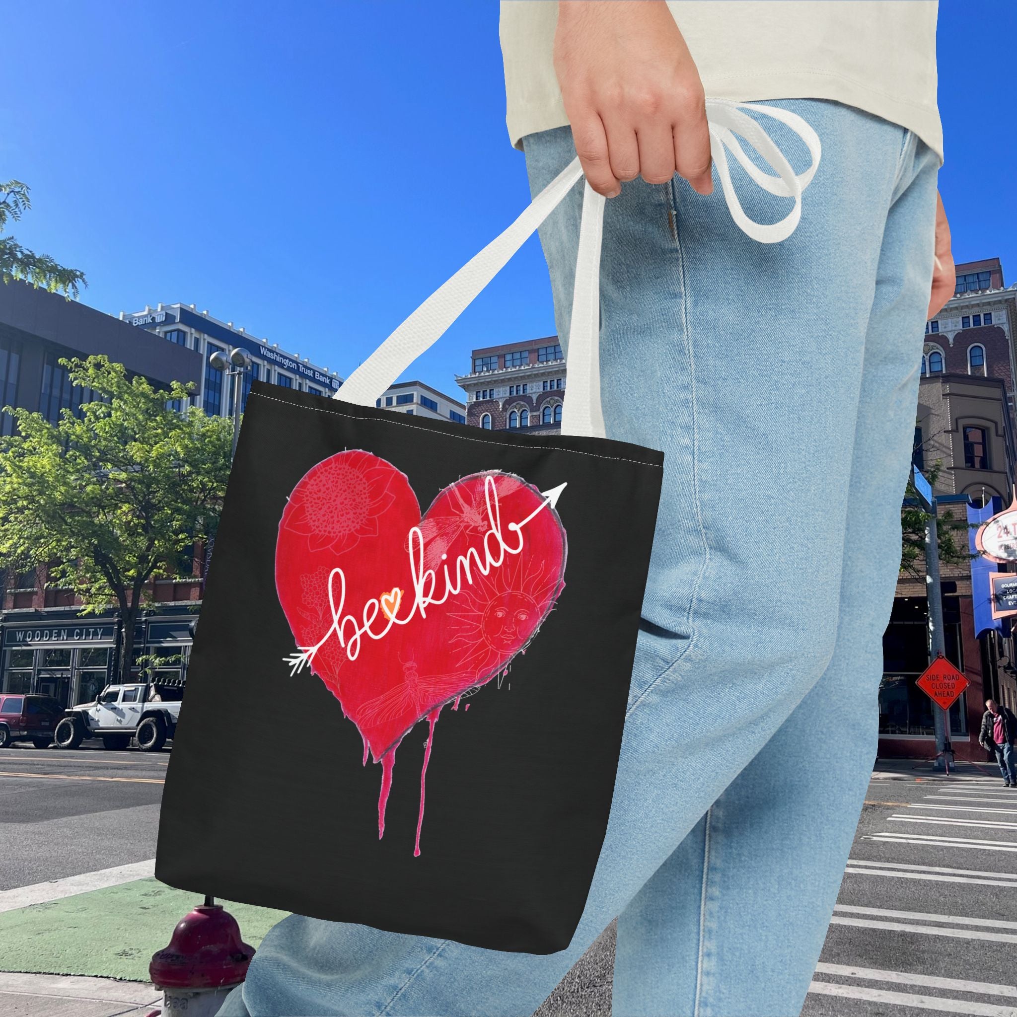 Person holding a black tote bag with a red heart and 'be kind' text on a city street.