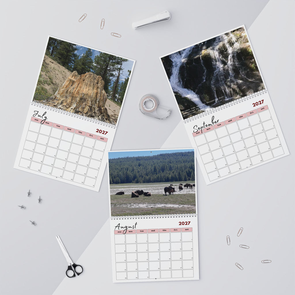 Three spiral-bound monthly desk calendars arranged on a light gray surface with small office props. Each calendar shows a different nature photograph: a rock formation in July, a waterfall in September, and bison in August.