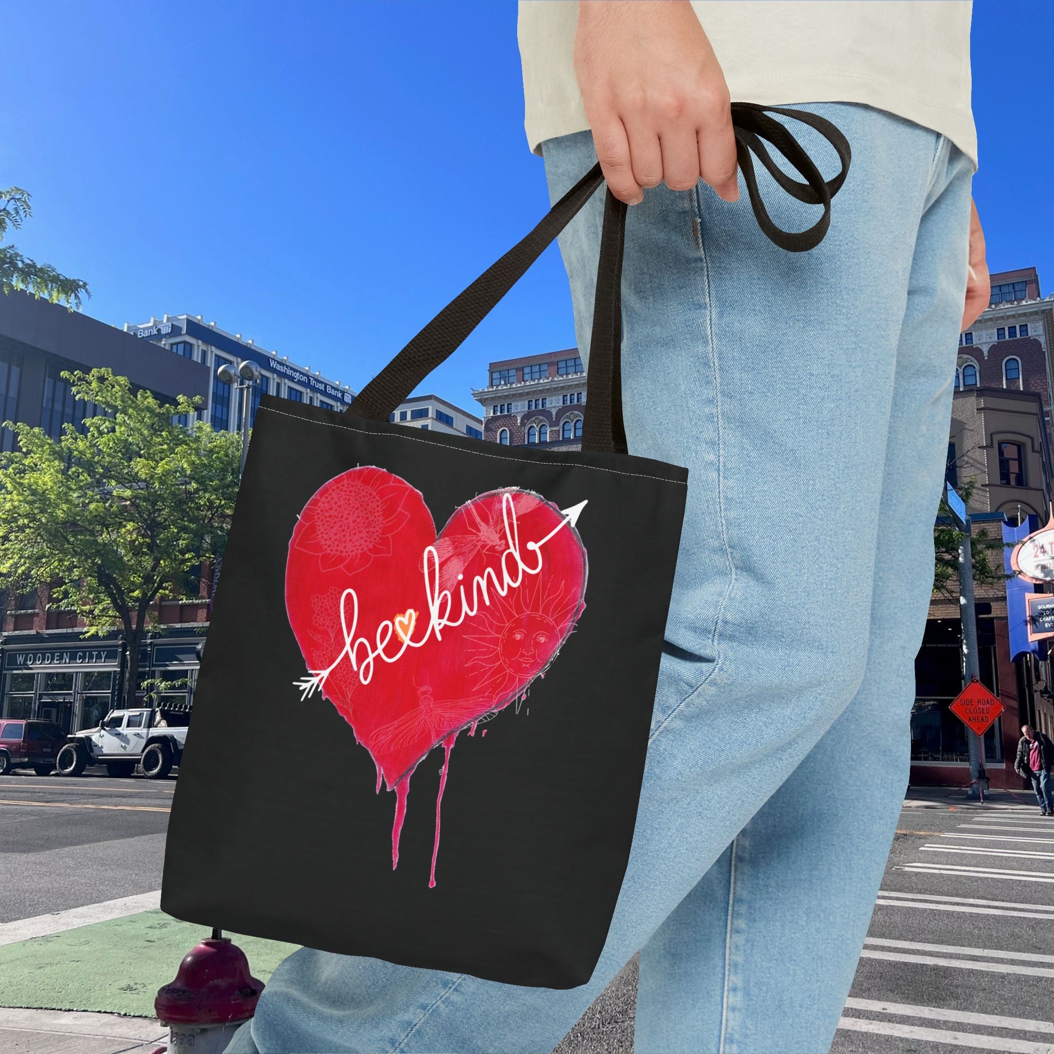 Person holding a black tote bag with a red heart and 'be kind' text on a city street.