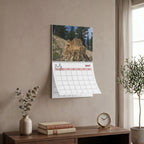 A spiral-bound wall calendar featuring a July 2027 page with a mountain landscape photo, hanging on a beige wall above a wooden console table with books, a vase, and a small clock.