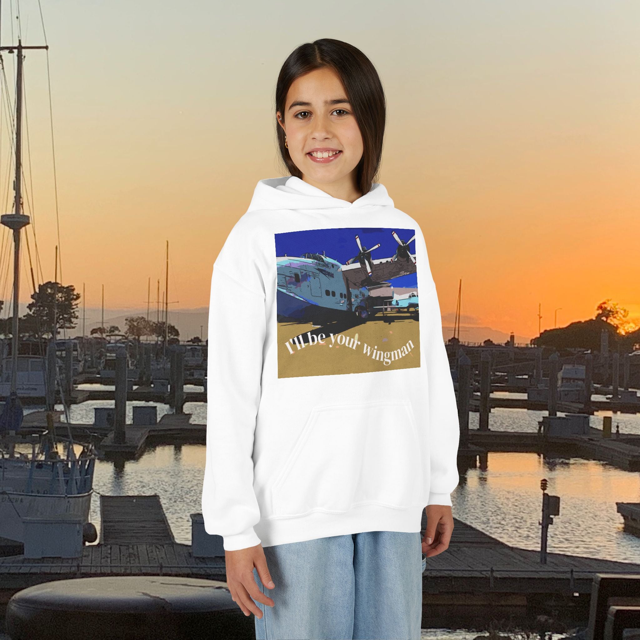 Person wearing a hoodie with an airplane graphic and text, standing in front of a marina at sunset.