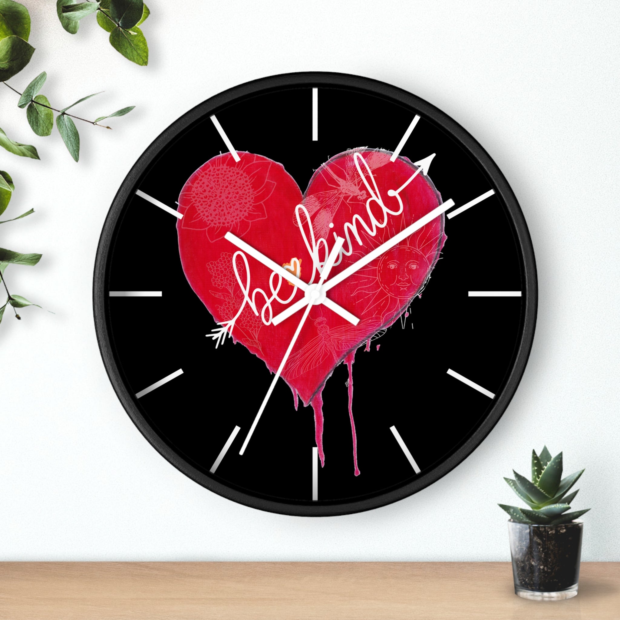 Wall clock with a red heart and 'be kind' text on a black background, placed on a wooden surface with a plant.
