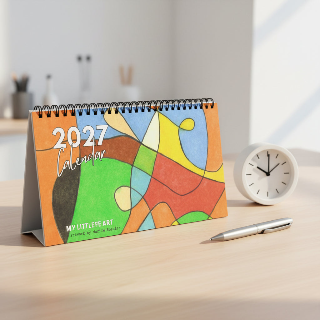 A 2027 spiral-bound desk calendar titled 'MY LITTLE FE ART' featuring an abstract multicolor art design on the cover, photographed on a desk with a pen and clock next to it.