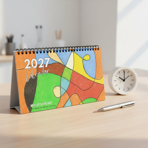 A 2027 spiral-bound desk calendar titled 'MY LITTLE FE ART' featuring an abstract multicolor art design on the cover, photographed on a desk with a pen and clock next to it.