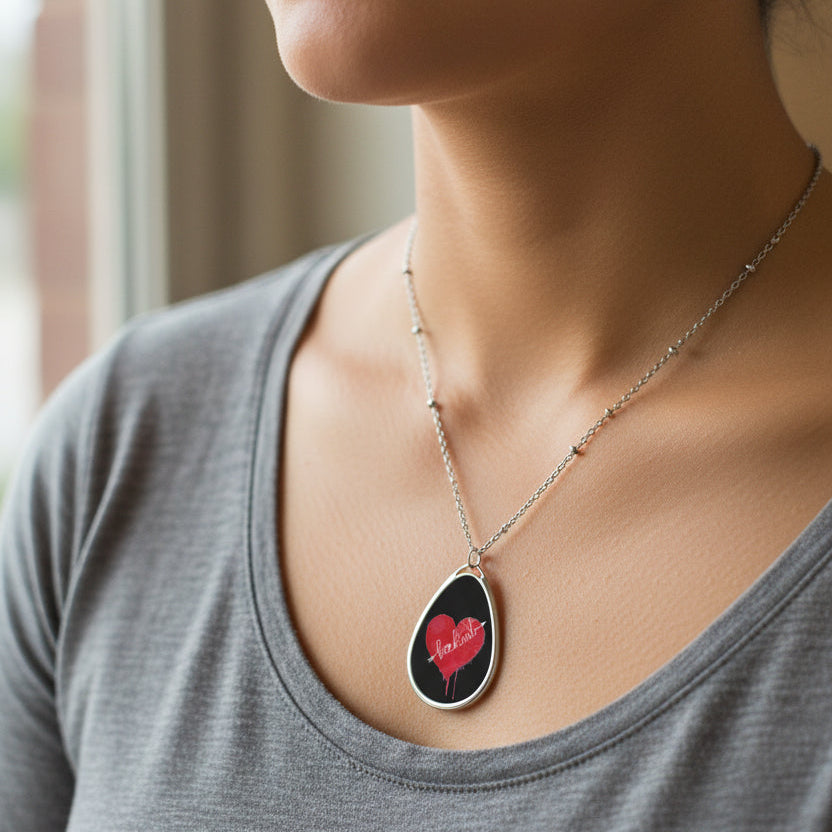 A silver teardrop pendant necklace featuring a black background with a red heart design and the word 'be kind' written across it on a woman's neck