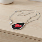 A silver teardrop-shaped pendant necklace featuring a black background with a red heart design and the word 'bookimb' in white script, displayed on a light wood dresser