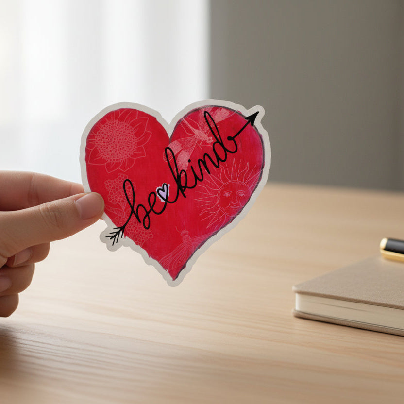 Hand holding a red heart-shaped sticker with 'be kind' text and decorative elements on a desk with a notebook next to it