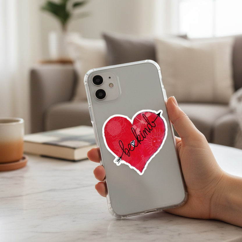 hand holding a phone cas with a red heart-shaped sticker with 'be kind' text and decorative elements on a coffee table with coffee, book and sofa in the background