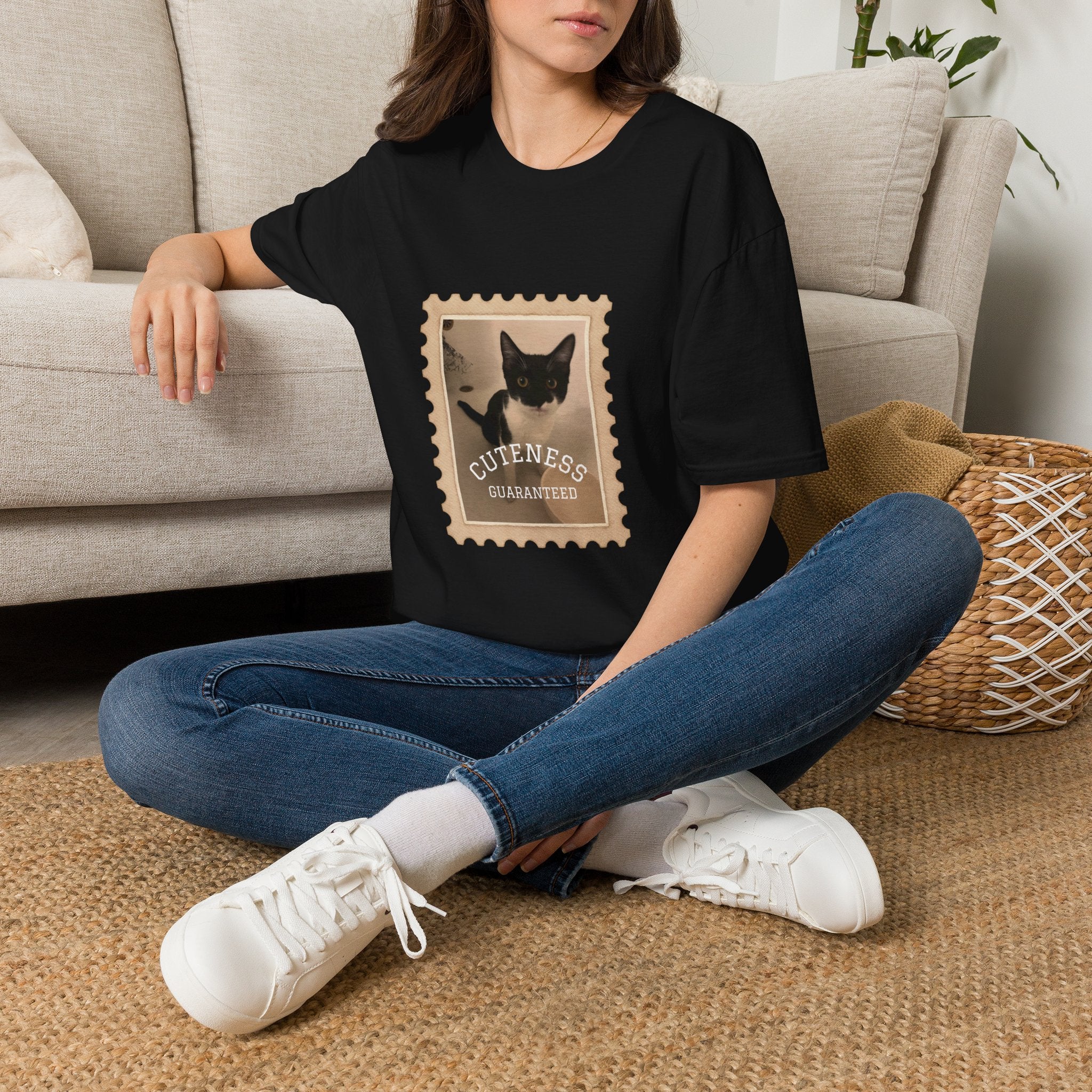 A woman sitting on a woven rug in front of a beige sofa, wearing a black oversized T-shirt with a vintage-style postage stamp graphic featuring a black-and-white cat and the words 'Cuteness Guaranteed.'.