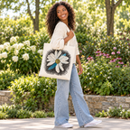 A smiling woman with curly hair carries a black canvas tote bag at an outdoor farmers market. The tote bag features a large white daisy graphic with a blue accent and black sketch-style detailing.