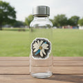 Sticker with a daisy dragonfly wings petal design on a claer water bottle on a picnic table in a park setting.