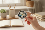 Daisy Dragonfly Wings Petals sticker held by a hand at a home office desk with a notebook, books a plant and pencils around it