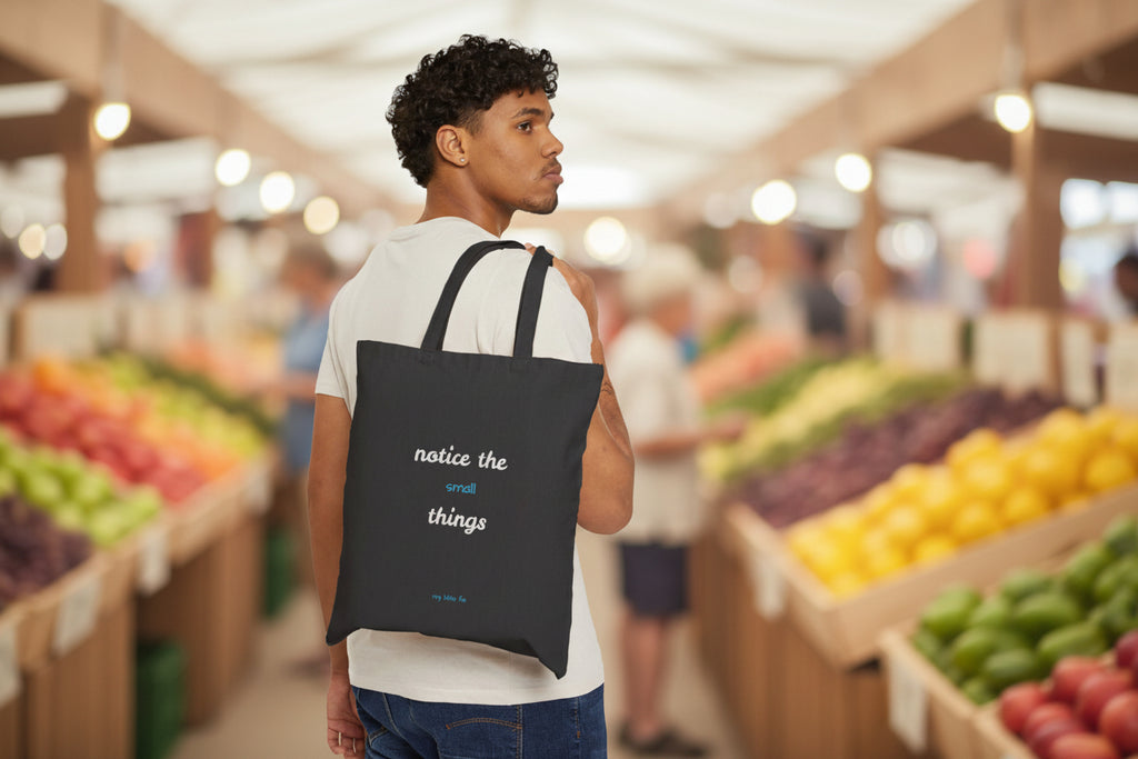Man carrying a black tote bag with text notice the small things at a farmer's market