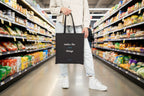 Person holding a black tote bag with text 'notice the small thngs' at an aisle in a grocery store