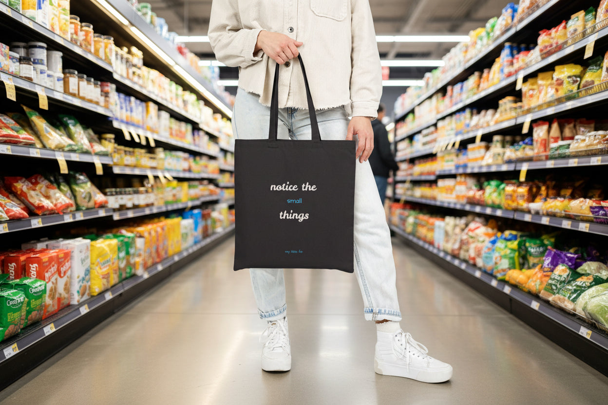 Person holding a black tote bag with text 'notice the small thngs' at an aisle in a grocery store