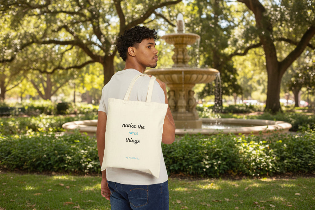 Man carrying a tote bag showing the back side with text notice the small things at a park with a fountain