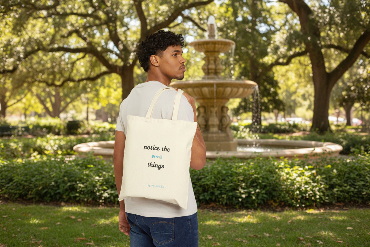 Man carrying a tote bag showing the back side with text notice the small things at a park with a fountain