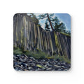 A square coaster with rounded corners featuring a painted landscape of a tall columnar cliff, green trees, and a blue sky, shown on a plain white background.