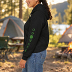 A woman shown from the side wearing a black cropped hoodie with green vertical text reading 'ODONATA' and a green circle on the sleeve, paired with blue jeans, against a plain white background.